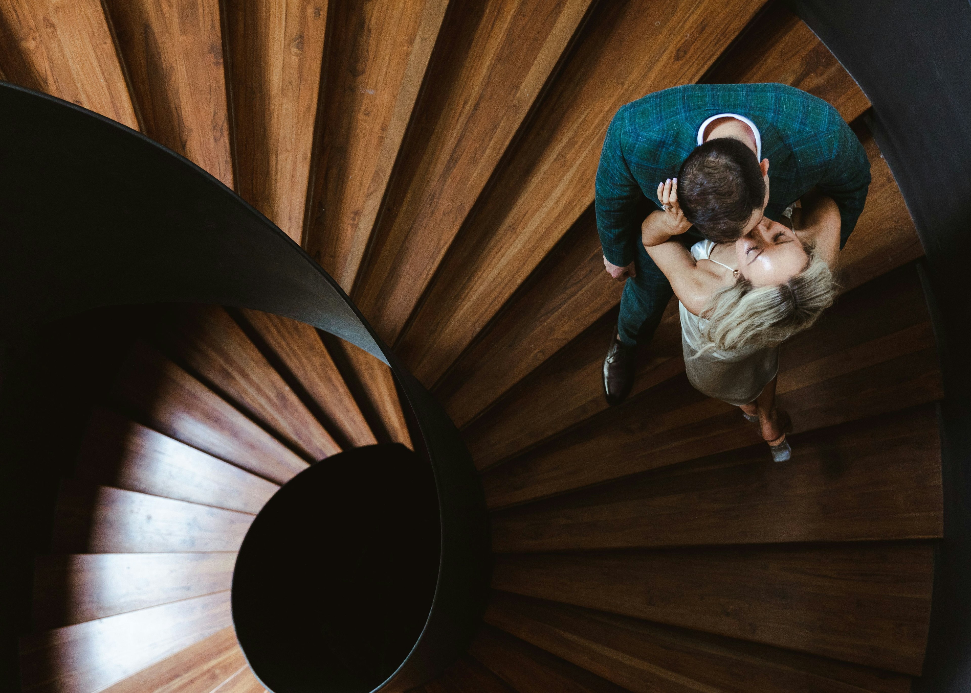 An aerial view of an intimate moment, capturing a couple sharing a kiss on a spiraling wooden staircase at Vivid Grand Island, the architecture framing their embrace in a stunning geometric composition.