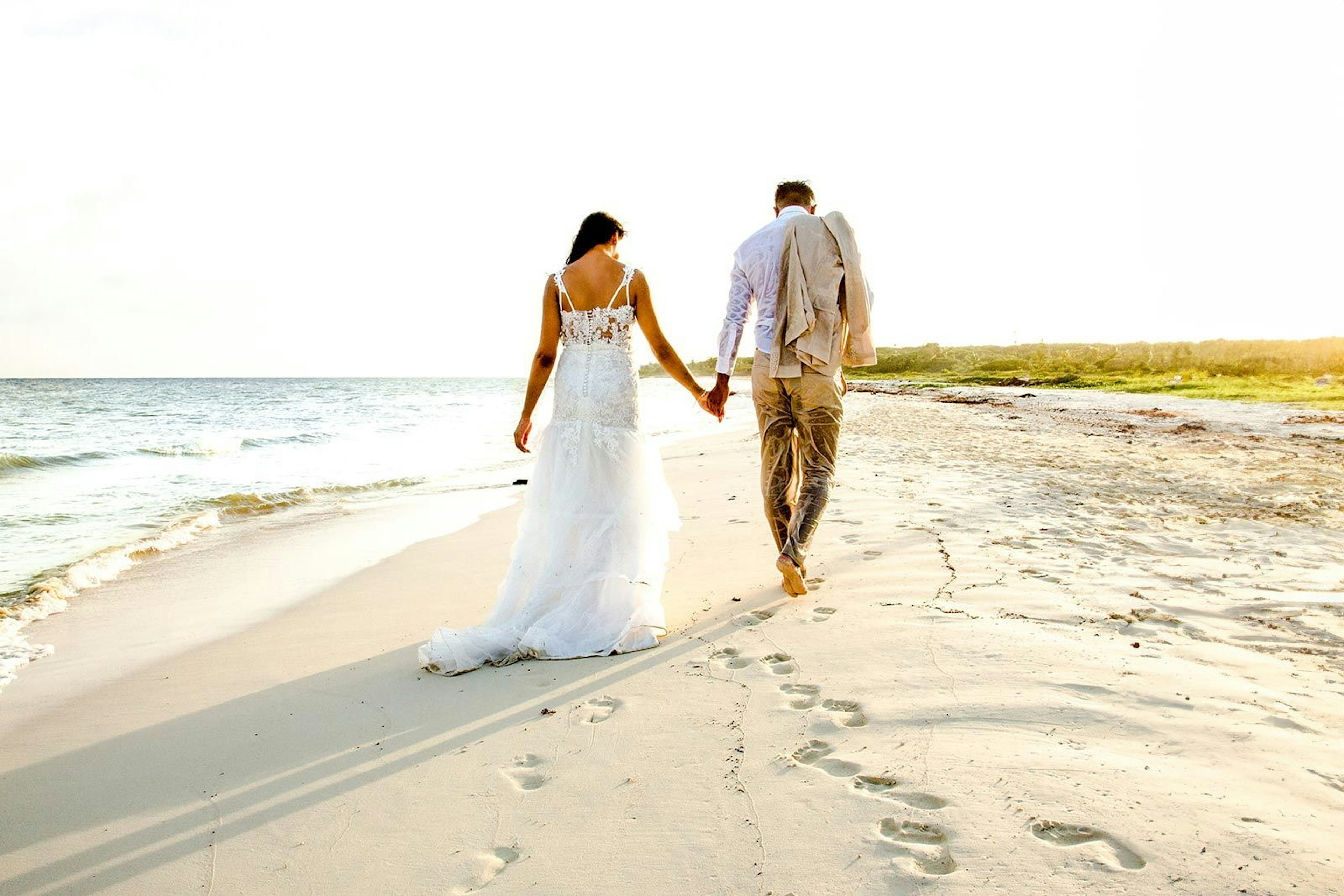 A couple strolls hand in hand along the beach at Secrets Tulum, leaving footprints in the sand as the setting sun casts a warm glow over their tranquil walk, the bride's elegant dress trailing behind.